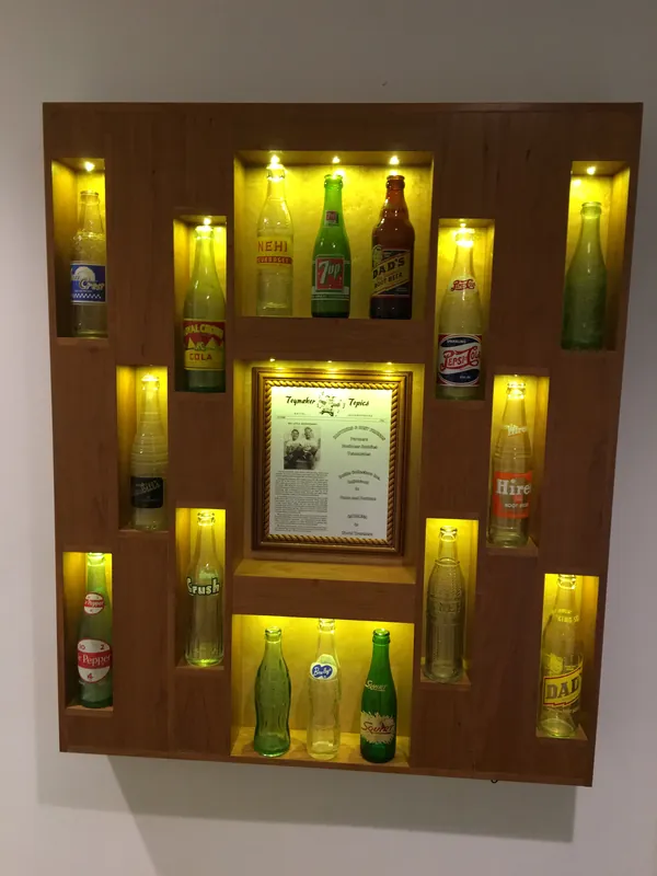 Finished cherry pop bottle display with lit bird’s-eye maple niches holding vintage soda bottles around a framed newsletter article, Agoura Hills