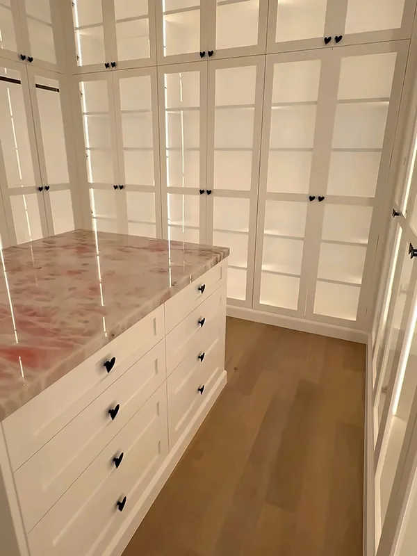 Walk-in closet with a white drawer island topped in pink marble, facing a wall of LED-lit glass-front cabinets